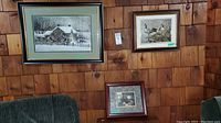 Three framed pictures hanging on wood-paneled wall including a large framed snowy house scene, a medium framed bird scene, and a smaller framed still life.