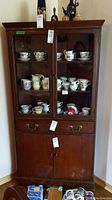 Front view of wood corner cabinet with glass doors showing shelves filled with ceramic cups and the drawer and wooden lower cabinet closed.