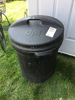 Black plastic garbage can with textured lid featuring a raised handle and embossed 'dpl' logo, placed on grass outdoors