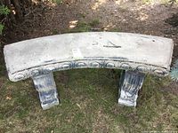 Front view of a curved cement garden bench with decorative circular carved patterns along the edge and sturdy legs, placed on grass outdoors.
