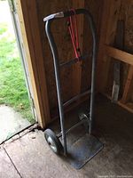 Full view of the metal hand cart showing the curved handle, rubber wheels, and flat metal platform. Red bungee cord attached to handle.