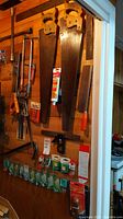 Wall-mounted display of several hand saws with wooden and plastic handles, assorted small hardware packets below.