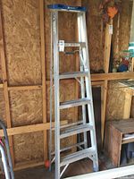 Full view of the 6 ft aluminum folding ladder standing upright against a wooden wall with a blue top platform and six wide steps.