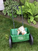 Shows full green Scotts AccuGreen 1000 push broadcast spreader with black handle and wheels placed on grass near plants.