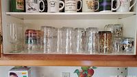 Shelf with assorted ceramic mugs on top row and various clear glass tumblers and goblets on lower row.