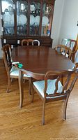 Photo showing wooden oval dining table with six chairs placed around it in a room with hardwood floors and a china cabinet in background.
