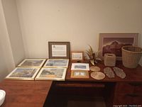 Four framed landscape pictures, large framed building picture, wicker baskets, pan holders, stone carvings and small decor items on wooden desk.