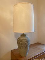Green ceramic style lamp with beige tall cylindrical fabric shade, placed on wood surface near wall.