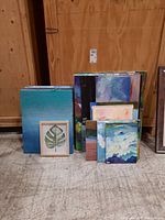 Group of over 15 paintings on canvas and board, stacked together showing various abstract and landscape styles, with one framed botanical leaf print visible in front
