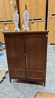 Front view of vintage solid wood cabinet showing two paneled doors with the key in place and lamps on top.