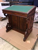 Photo showing front and top angled view of wooden desk base with green felt surface bordered by brass studs, ornate scrolling wood supports, and drawer fronts.