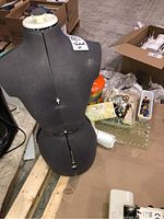 Gray adjustable dress form mannequin standing on wooden planks with boxes and containers of sewing supplies scattered around it on the floor.