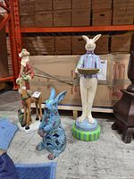 Photo showing tall standing bunny figurine in pastel plaid jacket holding basket alongside blue seated bunny figurine and additional unrelated figures in background.