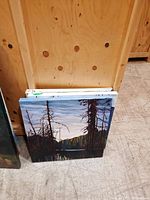 Three original landscape paintings on square canvases with bare trees and lake scene, stacked and against wooden background.