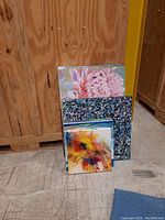 Three unframed paintings on canvas, stacked showing floral and abstract designs in colorful palettes.
