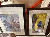 Two framed and matted paintings side by side: one of a moose with large antlers on a cosmic background, the other of a bicyclist on a sunny tree-lined path with a dog.