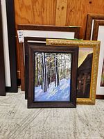 Photo showing multiple framed paintings, with a prominent view of a snowy forest painting with dark wooden frame and partially visible gold frame behind