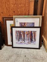 Three framed watercolour paintings leaning against a wall, showing forest and nature scenes.