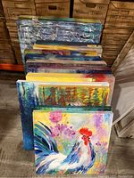 View of all twelve paintings stacked upright showing varied colorful painted canvases including a rooster, nature scenes, and abstract art.