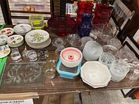 Photo showing multiple glass vases in red and blue, clear glass bowls and plates, glass juicers, ceramic plates with botanical prints, and glass paperweights arranged on a table.