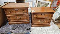 Two wooden side tables with two drawers each, medium brown finish, brass hardware, decorative wood grain and trim details, stacked on moving blankets in storage area.