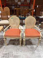 Front view of two matching vintage occasional chairs with painted wood frames, caned backs and orange upholstered seats.