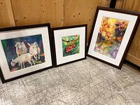 Photo of all three framed watercolour paintings arranged on floor. One large vertical painting with colorful trees and canoe, one medium square painting with three goats, one small square painting with cherries.