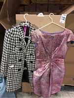 Houndstooth jacket and pink purple paisley dress hung on hangers against cardboard boxes.