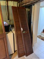 Two wooden bifold doors leaning against a washing machine and wall in a basement, showing full length and louvered design with minor scratches.