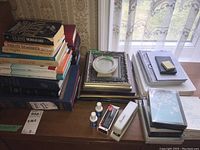 Photo showing stacks of books, picture frames, stationery items including notebooks and note cards, a white stapler, and small bottles on a wooden surface in front of a window.