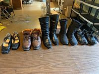 View of the full line-up of seven women's shoes and boots on floor, showing varying styles including boots and sneakers all in size 8.5