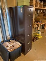Full height side view of dark brown metal filing cabinet with five drawers, placed near basement wall next to a large plastic container with wood pieces.