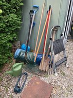 Overall photo showing assortment of rakes, shovels, watering can, step ladder and snow shovels standing on gravel surface near a bush and siding of a building.