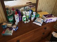 Photo of various health and personal grooming products arranged on a wooden dresser with a mirror in the background. Includes mouthwash, lotions, ointments, wipes, denture cleanser and personal hygiene products in retail packaging.