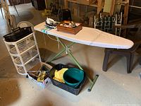 Photo showing the ironing board set up with sewing supplies on top and various other items around it including plastic cart and wire hangers.