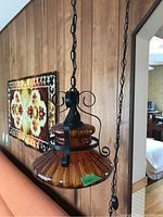 View of amber glass hanging lamp with black metal decorative scroll frame and hanging chain, mounted indoors against wood paneling.