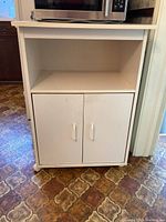 Front view of white melamine microwave stand with open top shelf and closed lower cabinet with two doors and plastic handles.