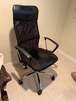 Black office chair with vinyl padded seat and headrest, mesh backrest, plastic armrests, and chrome metal base with caster wheels, placed on tiled floor beside furniture.