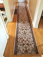 Long hallway runner carpet with intricate floral design in brown, beige, and cream tones shown laid out on hardwood floor.