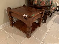 Wooden side table with a framed top, four turned legs, and a lower shelf, shown from an angle against a tiled floor.