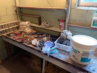 Wide view of basement workspace showing hand tools, hardware, plastic drawers, mini fans, bucket of masonry coating, and drop cloths on a workbench.