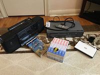Wide view showing Magnavox VHS/DVD player, Yorx portable stereo, D-Link router, VHS tapes and remote control on carpeted floor.