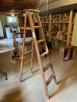 Wooden 6-foot step ladder standing in basement, visible paint stains and metal braces on structure, photo shows ladder from an angled side view.