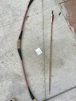 Wooden recurve bow with black handle grip and red limbs next to one wooden arrow with red and white fletching and pointed tip.