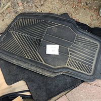 Black rubber floor mat with angular groove patterns on top of dark cloth mat on outdoor ground.