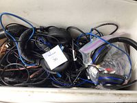 Close-up photo showing multiple used electrical cords, phone cables, and several wired headphones tangled inside a container.