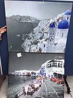 Photo showing the two framed scenic pictures, upper one displaying a coastal view with blue domes and cliffs, lower one showing a promenade with chairs and white buildings along the water.