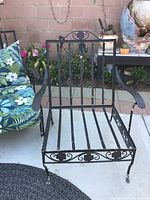 Front view of a black metal patio chair frame showing vertical slats, floral scrollwork on the back and base, and metal armrests; cushions are not included.