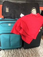 Photo showing two suitcases (teal and black), large black army duffel bag, and five red kid polo shirts on hangers.