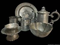 Photo showing six pewter items including a decorative plate, teapot, two tankards, and two bowls with a dark background.